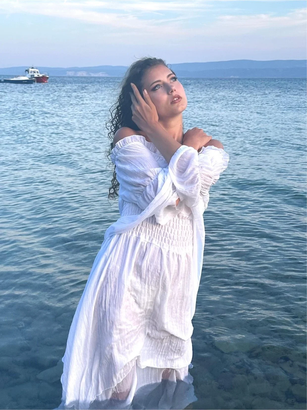 Samia Winkler standing in the sea in a white dress, eyes closed, hands at her face