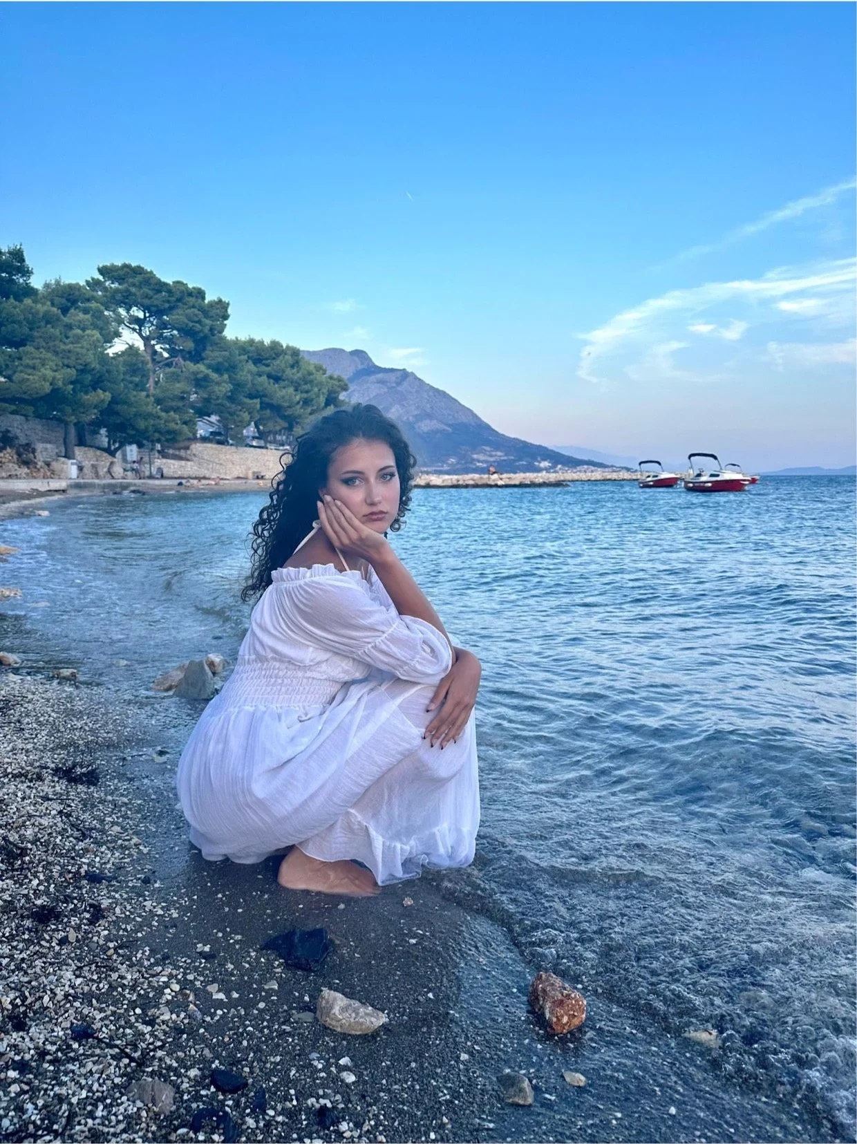 Samia Winkler crouching on a pebble shore in a white dress, mountains in the background