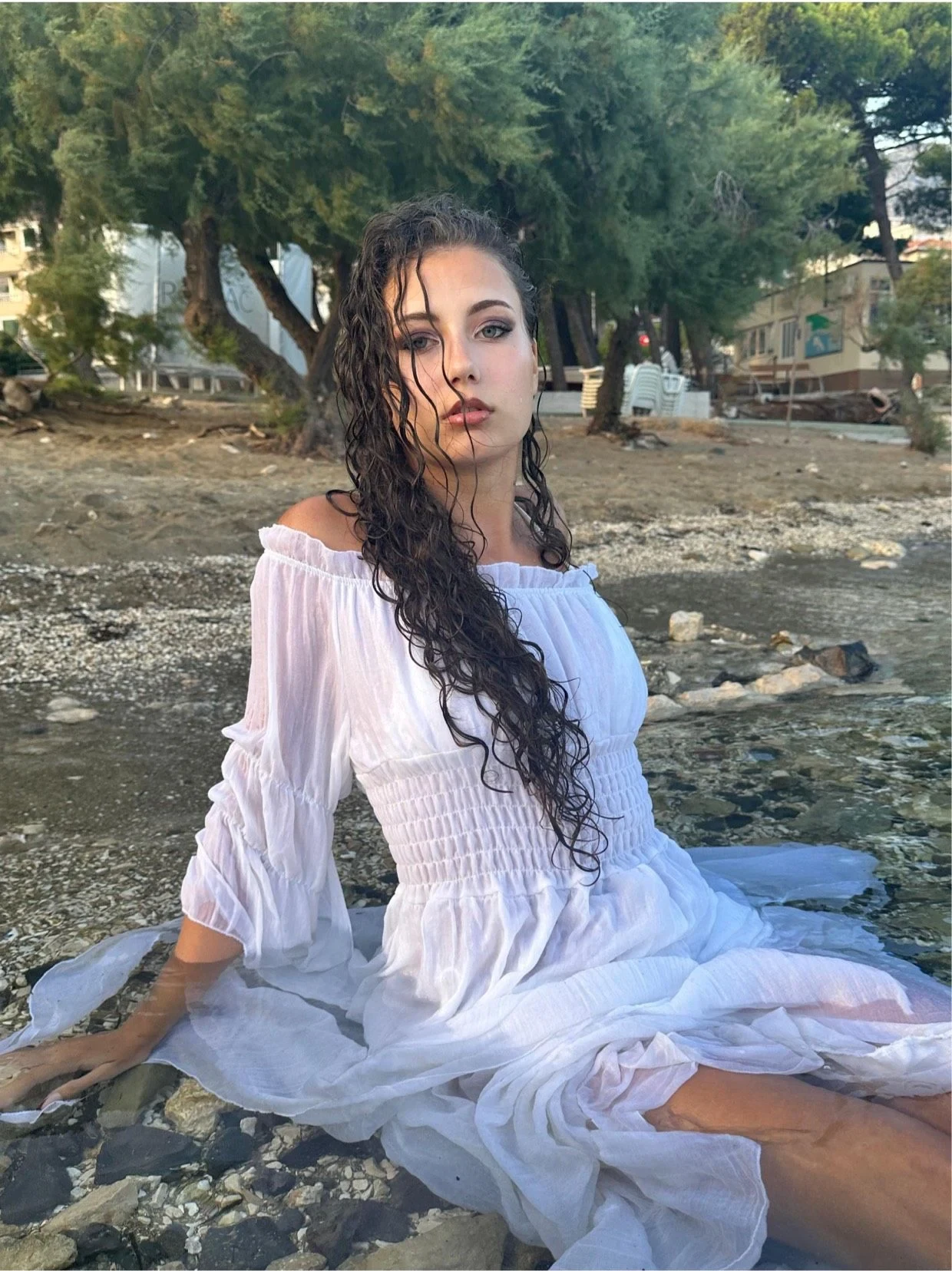 Samia Winkler sitting on a stony beach in a white off-shoulder dress, wet curly hair