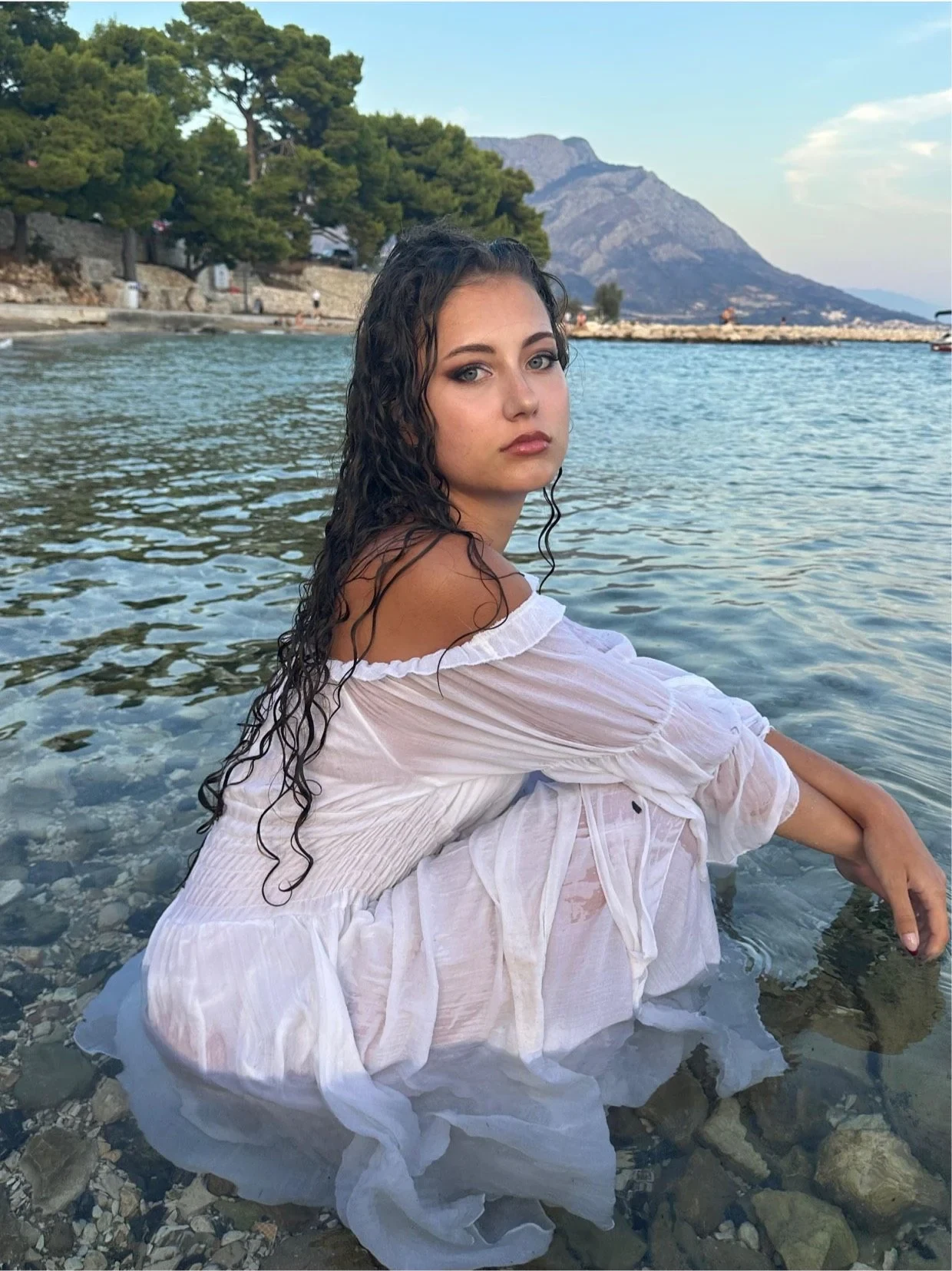 Samia Winkler kneeling in shallow water in a white off-shoulder dress, mountains behind